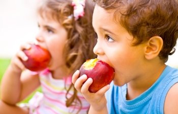 Photo of two happy children eating apples, brother and sister having picnic outdoors, cheerful kids biting red ripe peach, adorable infant holding fresh fruits in hands, healthy nutrition concept
