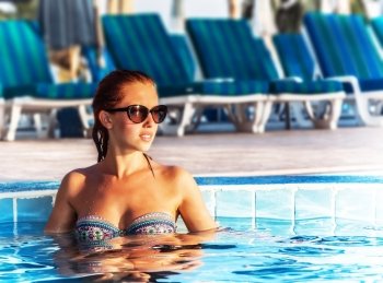 Image of beautiful woman in pool, pretty female taking sun bath in poolside, sexy girl wearing stylish sunglasses and colorful swimwear, luxury beach resort in hotel, tropical vacation, travel concept