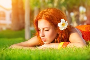 Portrait of cute calm girl lying down on fresh green grass with franjipani flower in red hair, dreamy closing eyes, enjoying day spa on luxury beach resort
