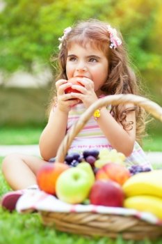Portrait of a cute little girl sitting on the green grass field in a park and with pleasure eating tasty juicy apple, healthy babies nutrition