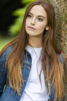Outdoor portrait of beautiful thoughtful sad girl or young woman with red hair wearing a white t-shirt and denim jeans jacket leaning against a tree in the countryside