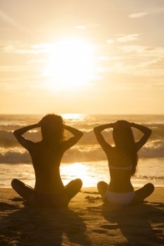 Two relaxed sexy young women or girls wearing bikinis sitting on a deserted tropical beach at sunset or sunrise