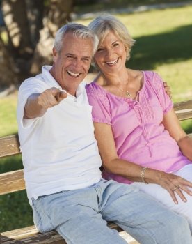Happy senior man and woman couple sitting together laughing and pointing on a park bench outside in sunshine