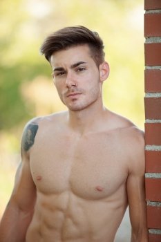 Handsome fit athletic shirtless young man with a tattoo leaning against a brick wall