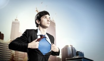 Young superhero businessman. Image of young businessman showing superhero suit underneath his shirt standing against city background