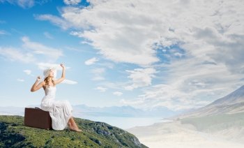 She is traveling light. Woman in white long dress and hat sitting on her luggage on mountain top