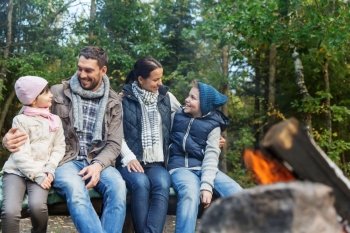 camping, travel, tourism, hike and people concept - happy family sitting on bench and talking at camp near campfire in woods