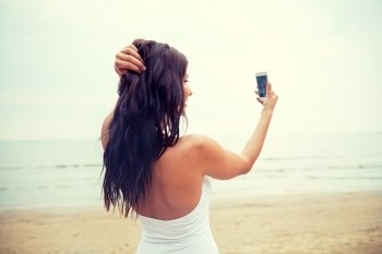 summer, travel, technology and people concept - sexy young woman taking selfie with smartphone on beach from back