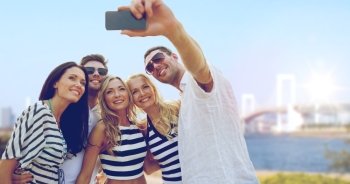 summer holidays, travel, tourism, technology and people concept - group of smiling friends with smartphone photographing and taking selfie over rainbow bridge in tokyo in japan background