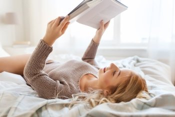 rest, comfort, leisure and people concept - happy young woman reading book in bed at home bedroom