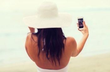 lifestyle, leisure, summer, technology and people concept - smiling young woman or teenage girl in sun hat taking selfie with smartphone on beach