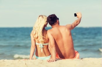 love, travel, tourism, technology and people concept - smiling couple on vacation in swimwear sitting on summer beach and taking selfie with smartphone from back