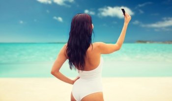 summer, travel, technology and people concept - sexy young woman taking selfie with smartphone over tropical beach background