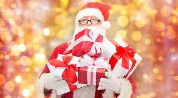 christmas, holidays and people concept - man in costume of santa claus with gift boxes over lights background