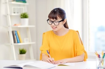 people, education, high school and learning concept - happy asian young woman student in glasses with book and notepad writing at home