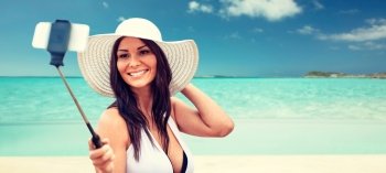 lifestyle, leisure, summer, technology and people concept - smiling young woman or teenage girl in sun hat taking picture with smartphone on selfie stick over tropical beach background