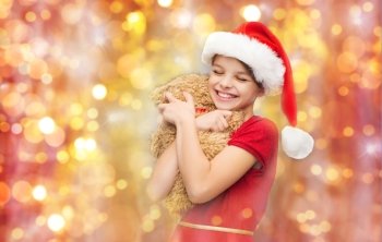 christmas, holidays, children and happiness concept - smiling girl in santa helper hat with teddy bear over lights background