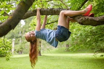friendship, childhood, leisure and people concept - happy smiling little redhead girl hanging upside down on tree in summer park