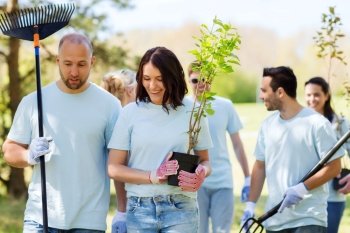 volunteering, charity, people and ecology concept - group of happy volunteers with tree seedlings and rake walking in park