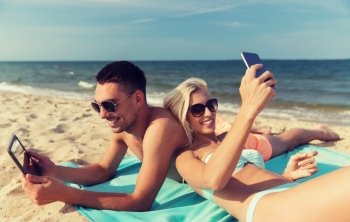 love, travel, tourism, technology and people concept - happy couple on vacation with tablet pc computer and smartphone sunbathing on summer beach