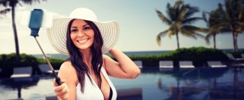 lifestyle, leisure, summer, technology and people concept - smiling young woman in sun hat taking picture with smartphone on selfie stick over resort beach with palms and swimming pool background