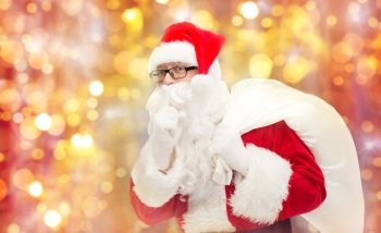 christmas, holidays and people concept - man in costume of santa claus with bag making hush gesture over lights background