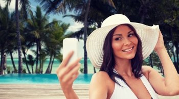travel, summer, technology and people concept - sexy young woman taking selfie with smartphone over tropical beach with palms and swimming pool background