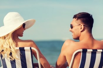 love, travel, tourism, summer and people concept - smiling couple on vacation in swimwear sitting in chairs and sunbathing on beach from back