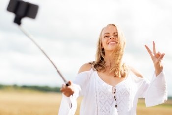 technology, summer holidays, vacation and people concept - smiling young woman in white dress taking picture by smartphone selfie stick and showing peace sign on cereal field