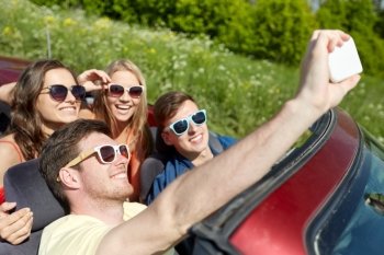 leisure, road trip, travel and people concept - happy friends driving in cabriolet taking selfie by smartphone at country