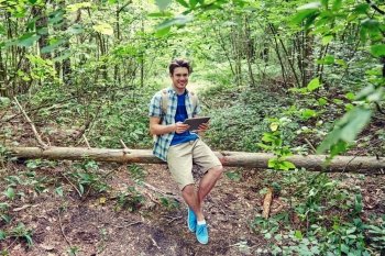 adventure, travel, tourism, hike and people concept - happy young man with backpack and tablet pc computer sitting on fallen tree trunk in woods