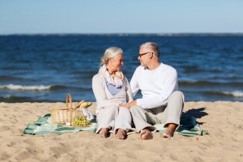 family, age, holidays, leisure and people concept - happy senior couple with picnic basket sitting on blanket on summer beach