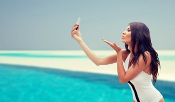 summer, travel, technology and people concept - sexy young woman taking selfie with smartphone and sending blow kiss over beach and swimming pool background