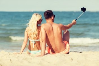 love, travel, tourism, technology and people concept - smiling couple on vacation in swimwear sitting on summer beach and taking picture with smartphone selfie stick from back