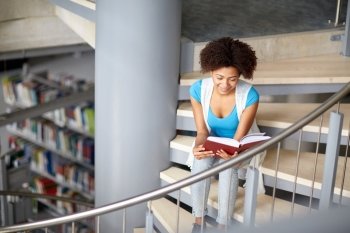 education, high school, university, learning and people concept - smiling african american student girl reading book sitting on stairs at library