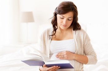 pregnancy, people and motherhood concept - pregnant woman reading book sitting on bed at home bedroom