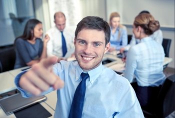 business, people, gesture and teamwork concept - smiling businessman pointing finger to you with group of businesspeople meeting in office