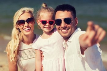 travel, vacation, adoption and people concept - happy family with little girl in sunglasses pointing finger on summer beach
