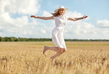 happiness, nature, summer, vacation and people concept - happy smiling young woman or teenage girl wearing straw hat and white dress jumping on cereal field
