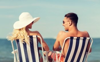 love, travel, tourism, summer and people concept - smiling couple on vacation in swimwear sitting in chairs and sunbathing on beach from back