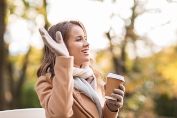 season, hot drinks and people concept - beautiful happy young woman drinking coffee or tea from disposable paper cup and waving hand in autumn park