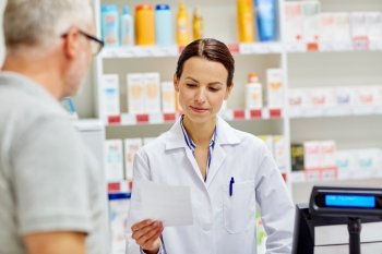 medicine, pharmaceutics, health care and people concept - pharmacist reading prescription and senior man at drugstore cash register
