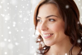 people, christmas and winter concept - close up of happy young woman looking through window at home over snow