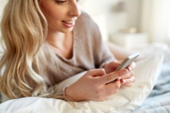technology, communication and people concept - close up of happy young woman lying in bed and texting on smartphone at home bedroom
