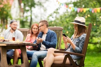 leisure, holidays, people and technology concept - young woman or teenage girl texting on smartphone and friends having dinner at summer garden party