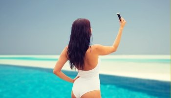 summer, travel, technology and people concept - sexy young woman taking selfie with smartphone over beach and swimming pool background