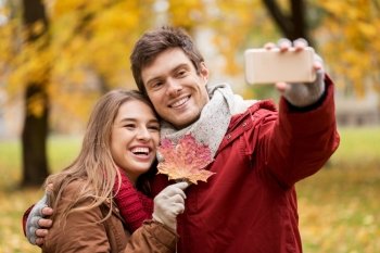 love, technology, relationship, family and people concept - smiling couple with maple leaf taking selfie by smartphone in autumn park