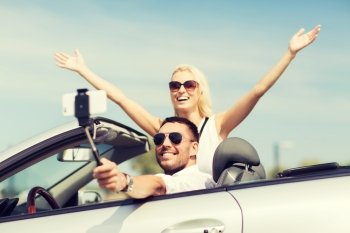 road trip, leisure, couple, technology and people concept - happy man and woman driving in cabriolet car and taking picture with smartphone on selfie stick