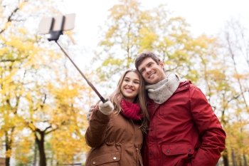 love, technology, relationship, family and people concept - happy smiling couple taking picture by smartphone selfie stick in autumn park. couple taking selfie by smartphone in autumn park