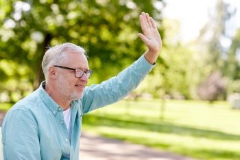 old age, gesture and people concept - happy senior man in glasses waving hand at summer park